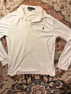 Vit långärmad piké från Polo Ralph Lauren - Klassisk vit långärmad pikétröja från Polo Ralph Lauren med blå broderad logga på bröstet. Tröjan har krage, knappar vid halsen och ribbade muddar vid ärmsluten. Tillverkad i mjukt bomullsmaterial som känns skönt mot huden.