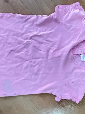 Rosa kortärmad t-shirt storlek 86 - Söt rosa t-shirt i bomull med korta ärmar och rund hals. T-shirten har knappar vid axeln för en extra detalj och en bekväm passform. Perfekt för en avslappnad och enkel stil.