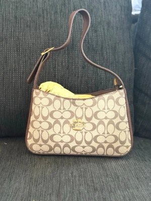 Beige axelremsväska från Coach - Snygg beige axelremsväska från Coach med klassiskt monogrammönster och bruna detaljer. Väskan har justerbart axelband, guldfärgad logga framtill och dragkedja upptill. Perfekt storlek för det viktigaste och ger en trendig touch till din outfit.