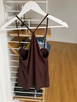 Brun ribbad crop top med smala band - Säljer en brun ribbad crop top med tunna axelband som korsas i ryggen. Toppen har en mjuk, stretchig känsla och är perfekt för lager på lager eller till jeans. Materialet är ribbat och ger en snygg struktur. Enkel och stilren design.