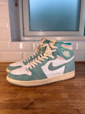 Air Jordan 1 High OG Turbo Green - Snygga Air Jordan 1 High OG Turbo Green sneakers med vit läderbas och turkosa / ljusblåa mockadetaljer. Klassisk hög siluett, vita skosnören och retro vingslogo på sidan. Storlek 44 / US10.