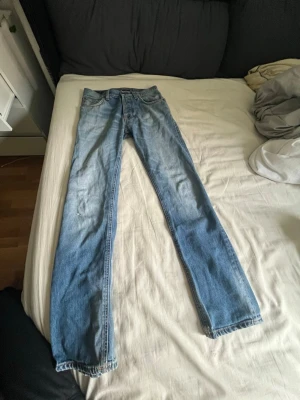 Blå raka jeans från Nudie Jeans - Säljer ett par klassiska blå jeans från Nudie Jeans i modellen med raka ben. Jeansen har en snygg tvätt med ljusare partier framtill och detaljer i orange söm. Perfekta för en avslappnad och stilren look.