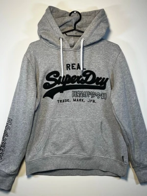 Grå hoodie från Superdry - Snygg grå hoodie från Superdry med stor svart logga och japansk text på bröstet. Hoodien har huva med vita snören, känguruficka och tryck på ena ärmen. Tillverkad i mjukt sweatshirtmaterial, perfekt för en avslappnad streetwear-look.