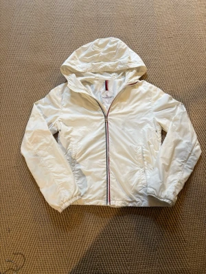 Moncler vindjacka – storlek 14 år - Snygg och stilren vindjacka från Moncler i storlek 14. Perfekt för vår, sommar och höst. Jackan är lätt och smidig med huva, dragkedja framtill och två praktiska fickor.  📏 Storlek: 14 år 🎨 Färg: Vit 🧥 Märke: Moncler ✨ Skick: Fint skick, se bilder för detaljer  En klassisk Moncler-jacka som passar till både vardag och en mer dressad outfit. Hör gärna av dig vid frågor!