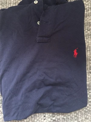 Marinblå pikétröja från Ralph Lauren - Klassisk marinblå pikétröja från Ralph Lauren med röd broderad logga på bröstet. Tröjan har krage, knappar framtill och är tillverkad i bomull. Custom fit-modell som sitter snyggt och passar till det mesta. (Pris går självklart att diskuteras😊)