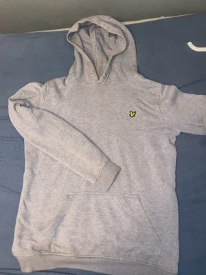 Ljusgrå hoodie med gul logga - Säljer en ljusgrå hoodie med huva och magficka. På bröstet sitter en liten gul logga som detalj. Hoodien har ribbade muddar vid ärmslut och nederkant. Perfekt för en avslappnad och stilren look.