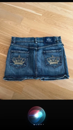 Blå jeanskjol med broderade fickor S/M - Snygg blå jeanskjol från Victoria Beckman med guldiga broderade kronor på bakfickorna. Klassisk femficksmodell med knapp och dragkedja framtill. Rå kant nedtill för en cool look. Perfekt för dig som gillar streetstyle.  Försäljaren sålde den som XS, men jag såg just att den faktiskt är en medium, vilket är anledningen till att den är så stor för min dotters storlek. Jag frågade till och med om den skulle passa en storlek 152/158, och hon sa ja, eftersom det var en liten modell. Passar bättre S
