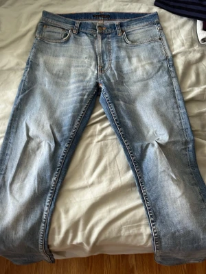 Nudie Lean Dean jeans - Säljer ett par ljusblå jeans i slim fit. Jeansen har klassisk femficksmodell, orangea sömmar och detaljer på bakfickorna. Perfekta för en avslappnad och trendig look. Har lagats på ett litet ställe på höger lår som knappt syns.