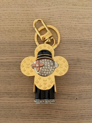 Vuitton guldfärgad nyckelring - Unik nyckelring från Vuitton i guldfärgad metall med ikoniskt LV-mönster. Figuren har svarta och silvriga detaljer, gnistrande stenar och en rosa blomdetalj. Perfekt som accessoar till väskan eller nycklarna.