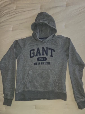 Grå hoodie från GANT med tryck - Snygg grå hoodie från GANT med mörkblå text 'GANT 1949 NEW HAVEN' på bröstet. Klassisk känguruficka framtill, ribbade muddar och huva. Tillverkad i mjukt bomullsmaterial som är perfekt för chill dagar.