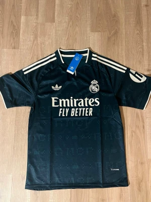 Real Madrid Bortatröja 26/27 Mörkgrön - Snygg svart Real Madrid bortatröja med vita detaljer och klubbmärke på bröstet. Tröjan har korta ärmar, rund hals och klassiska ränder på axlarna. Tillverkad i lätt och ventilerande funktionsmaterial, perfekt för fotboll eller träning. HELT NY!