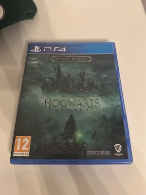 Hogwarts Legacy Deluxe Edition PS4 - Säljer spelet Hogwarts Legacy Deluxe Edition till PS4. Omslaget är mörkt med Hogwarts-slottet i bakgrunden och magiska detaljer. Spelet är i fysisk form med blå plastfodral och PEGI 12-märkning. Perfekt för dig som gillar äventyr och magi i Harry Potter-världen.