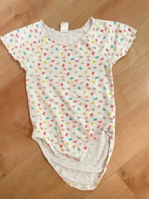 Vit body med färgglada fjärilar, H&M,86 - Supersöt vit body med korta ärmar och tryckknappar i grenen. Tunt och mjukt material med gulligt mönster av små fjärilar i rosa, gult, turkost och grått. Perfekt till sommaren och enkel att matcha med andra plagg.