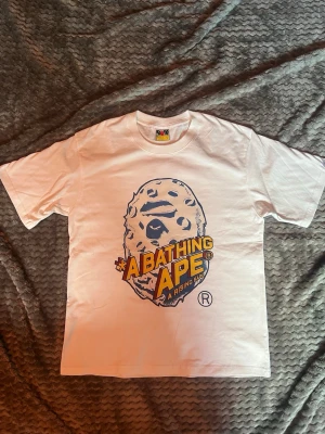 Vit BAPE t-shirt med stor logga - Säljer en vit t-shirt från A Bathing Ape med stor gul och svart logga och ikoniskt apansikte på bröstet. Klassisk passform och korta ärmar. Liten BAPE-tag på nederkanten. Perfekt för dig som gillar streetwear och vill sticka ut.