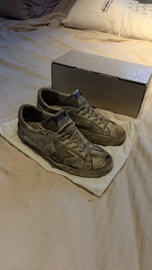 Beige sneakers med blå detalj - Säljer ett par beige sneakers från Golden Goose med blå logga på plösen och stjärndetalj på sidan. Skorna har en klassisk låg siluett, snörning och slitna detaljer för en vintage look. Tillverkade i mocka och läder med gummisula.