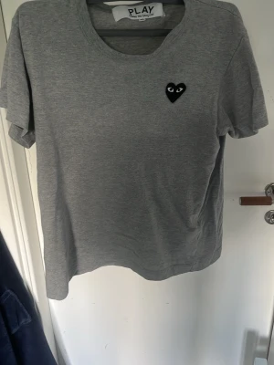 Grå Comme des Garçons PLAY t-shirt - Säljer en grå t-shirt från Comme des Garçons PLAY med det ikoniska svarta hjärta-märket på bröstet. T-shirten har rund halsringning och korta ärmar. Tillverkad i mjuk bomull som är skön att bära hela dagen. Storleken är xs och passar nån runt 165
