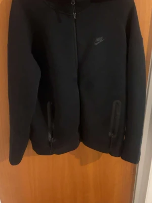 Svart Nike tech hoodie med dragkedja - Säljer en svart hoodie från Nike med hel dragkedja framtill och två fickor med dragkedja. Diskret Nike-logga på bröstet. Hoodien har en clean och sportig look, perfekt för dig som gillar streetwear. Materialet känns mjukt och bekvämt.