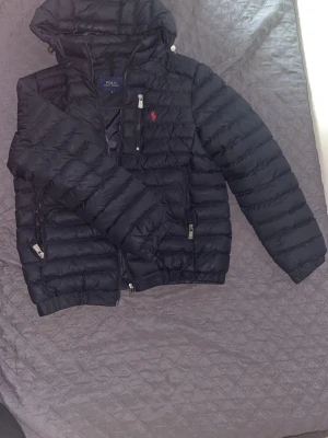 Svart pufferjacka från Polo Ralph Lauren - Snygg svart pufferjacka från Polo Ralph Lauren med röd broderad logga på bröstet. Jackan har huva, dragkedjor i silverfärg och två sidofickor med dragkedja. Perfekt för kalla vinterdagar och riktigt stilren look.