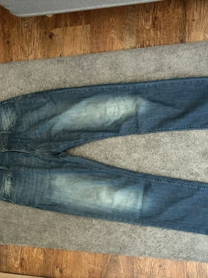 Blå jeans från Colin's Jeanswear - Säljer ett par blå jeans från Colin's Jeanswear med snyggt slitna detaljer och klassisk femficksmodell. Jeansen har en rak passform och är tillverkade i denim med ljusa tvättade partier på låren och knäna. Perfekta för en avslappnad stil.