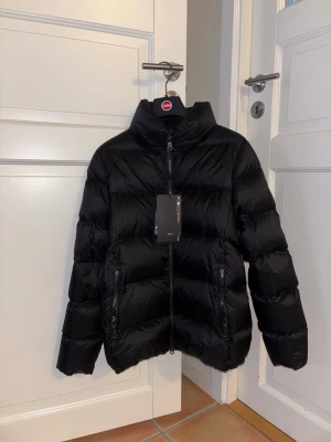 Svart dunjacka från Colmar - Snygg svart dunjacka från Colmar med hög krage och två dragkedjefickor. Jackan har en stilren design och är perfekt för kalla vinterdagar. Klassisk puffer-modell som håller dig varm och trendig. 14år passar mig som har xs ❤️