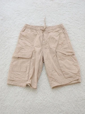 Beige cargoshorts med snörning - Säljer ett par beige cargoshorts med flera praktiska fickor och snörning i midjan. Shortsen har en avslappnad passform och är perfekta för varma dagar. Tillverkade i ett lätt och bekvämt material.