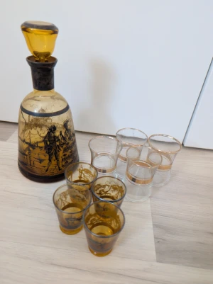 Hem tillbehör, paket - Säljer en vintage karaff med matchande snapsglas i bärnstensfärgat och klart glas. Karaffen har ett motiv av en stad och båtar, samt en dekorativ stopp. Totalt åtta glas, fyra i bärnstensfärg och fyra klara, alla med gulddetaljer. Perfekt för samlare eller som dekoration. Jag säljer även 5 glas skålar.
