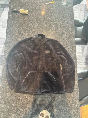 Svart teddyjacka från Reebok - Mysig svart teddyjacka från Reebok med dragkedja framtill och två stora fickor. Jackan har hög krage och är gjord i fluffigt teddy-material som håller dig varm. Diskret Reebok-logga på bröstet för en sportig touch. Finns ett hål till vänster om luvan vid axeln 