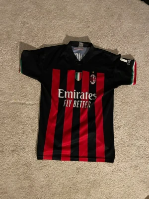 AC Milan matchtröja svart/röd - Snygg AC Milan matchtröja i svart och rött med klassiska vertikala ränder. Tröjan har klubbmärke och stjärna på bröstet samt sponsortryck 'Emirates Fly Better' framtill. Italienska flaggdetaljer på ärmarna. Perfekt för fotbollsfans!