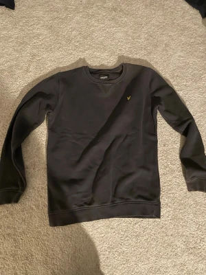 Svart sweatshirt från Lyle & Scott - Klassisk svart sweatshirt från Lyle & Scott Junior med rund halsringning och gul broderad logga på bröstet. Tröjan har ribbade muddar vid ärmslut och nederkant, perfekt för en avslappnad stil.