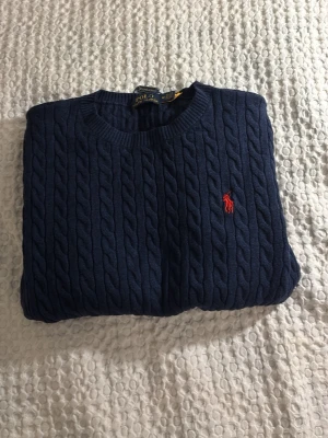 Mörkblå kabelstickad tröja Polo Ralph Lauren - Kabelstickad mörkblå tröja från Polo Ralph Lauren med klassisk röd broderad logga på bröstet. Tröjan har rund halsringning och ribbade muddar. Perfekt för dig som gillar tidlös och stilren design.