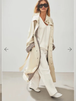 Beige trenchcoat med rutigt foder - Helt ny stilren beige trenchcoat med klassiskt rutigt foder på insidan. Lång modell med bälte i midjan och stora knappar framtill. Oversized passform och bred krage ger en cool vibe. Perfekt för lager-på-lager look. Nypris 1300kr