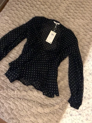 Svart prickig blus från ONLY - Snygg svart blus från ONLY med vita små prickar. Blusen har lång ärm, v-ringning och är gjord i ett tunt, transparent material som ger en luftig känsla. Perfekt för att skapa en trendig och stilren look.