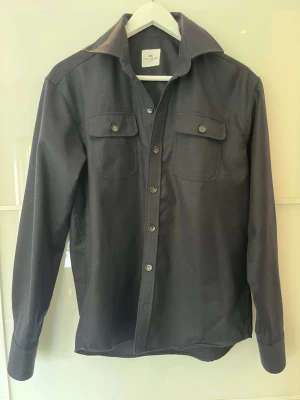 Svart overshirt från John Henric - Stilren svart overshirt från John Henric med långa ärmar och två bröstfickor med knappar. Den har klassisk krage och knäppning framtill. Perfekt för dig som gillar en enkel men snygg look.
