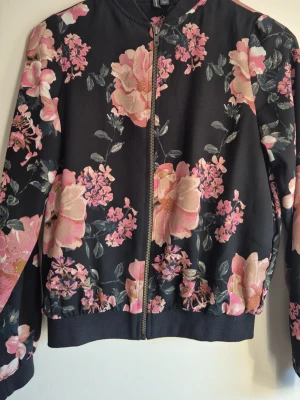 Blommig bomberjacka från Vero Moda - Snygg bomberjacka från Vero Moda i svart med stora rosa och ljusrosa blommor. Jackan har dragkedja framtill, ribbade muddar och en avslappnad passform. Perfekt för dig som vill sticka ut med ett färgstarkt mönster.