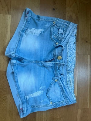 Ljusblå jeansshorts med rosetter - Säljer ett par ljusblå jeansshorts med hål och snygga rosettdetaljer på både fram- och bakfickor. Shortsen har gylf med dragkedja och knapp, samt dekorativa sömmar och bälteshällor. Bara att skriva om det finns nån fråga!💕