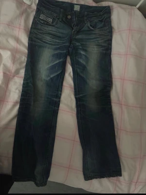 Mörkblå low waisted jeans med nitar - Säljer ett par mörkblå jeans med snygga slitningar och coola nitar på ena fickan. Jeansen har klassisk femficksmodell och är bootcut. De är väldigt lågmidjade och kommer passa dig i längden om du är mellan 154-162 cm lång. Storlek Xs men passar även S