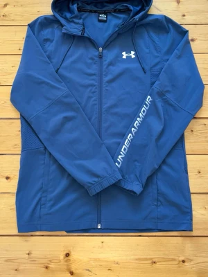 Grå vindjacka från Under Armour - Snygg grå vindjacka från Under Armour med huva och dragkedja framtill. Jackan har vitt logotryck på bröstet och UNDER ARMOUR-text på ena ärmen. Tillverkad i lätt och vindtätt material, perfekt för aktiva dagar utomhus.