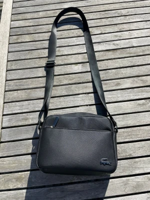 Svart axelväska från Lacoste - Snygg svart axelväska från Lacoste med justerbar axelrem och diskret krokodillogo framtill. Väskan har en slimmad, rektangulär form och är tillverkad i tåligt syntetmaterial med struktur. Utrustad med dragkedja både fram och bak för smidig förvaring.