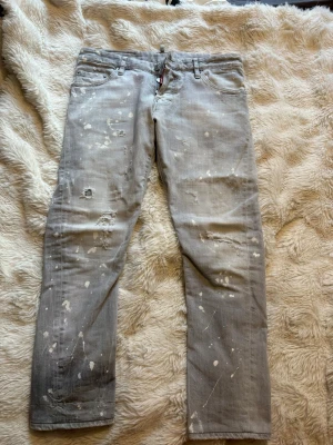 Grå distressed jeans från Dsquared2 resaverade - Säljer ett par grå jeans från Dsquared2 med coolt distressed utseende, slitningar och vita färgstänk över hela byxorna. Klassisk femficksmodell med raka ben och patch bak i midjan.  
