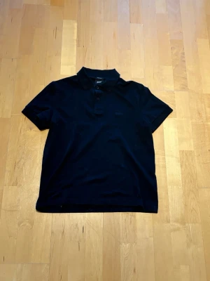 Svart kortärmad Hugo boss - Klassisk svart hugoboss piké med krage och knappar framtill. T-shirten har korta ärmar och en enkel, stilren design som passar till det mesta. Perfekt för dig som gillar en clean och tidlös look.
