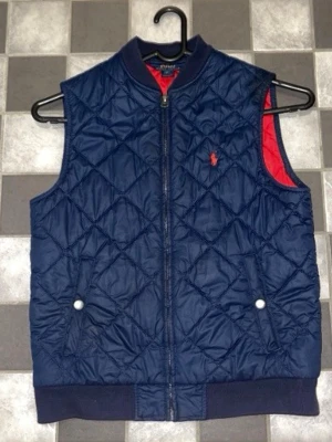 Mörkblå quiltad dunväst Polo Ralph Lauren - Snygg mörkblå dunväst från Polo Ralph Lauren med quiltat mönster och röd insida. Västen har dragkedja framtill, två fickor med tryckknappar och ribbade muddar nedtill. Perfekt för vår, höst och sommar väder