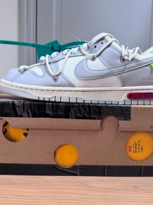 Deadstock Nike Off-White Dunk Low vit/grå - Oanvända Nike Off-White Dunk Low sneakers i vitt och ljusgrått läder och textil. Karaktäristiska detaljer som extra snörning, grön tag och rosa accent på sulan. Swoosh-logga på sidan och Off-White text på hälen. Kommer med originalkartong.