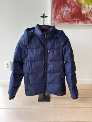 Mörkblå pufferjacka från Ralph Lauren - Snygg mörkblå pufferjacka från Ralph Lauren med röd broderad logga på bröstet. Jackan har avtagbar huva, två dragkedjefickor och grova sömmar för extra puffig look. Perfekt för kalla vinterdagar och riktigt varm.