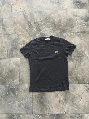 Svart t-shirt med Moncler-logga - Snygg svart t-shirt med Moncler-logga på bröstet. Klassisk rund halsringning och korta ärmar. Tillverkad i mjuk bomull för en skön känsla. Perfekt till jeans eller shorts för en clean look.