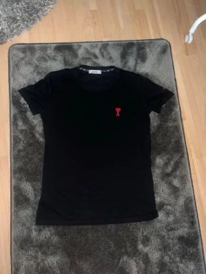 Svart t-shirt från AMI med rött hjärta - Svart t-shirt från AMI med ett litet rött hjärta broderat på bröstet och AMI-logga i rött på ärmen. Klassisk passform och rund halsringning. Perfekt till jeans eller shorts för en clean och stilren look.