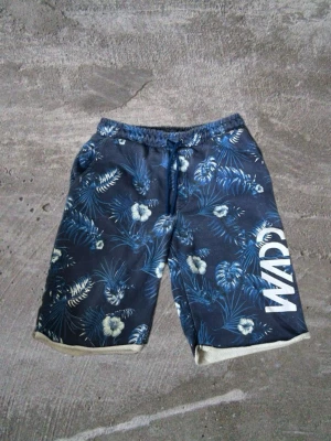 Blå  shorts med snörning - Snygga blå shorts med tropiskt blommönster och vita detaljer. Shortsen har resårmidja med snörning och tryckt text på ena benet. Perfekta för sommardagar och avslappnad stil.