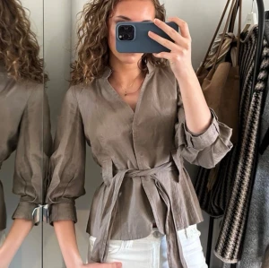 Beige blus med knytband i midjan - Snygg beige blus med trekvartsärmar och knytband i midjan som ger en feminin siluett. Blusen har v-ringad hals och lätt puffade ärmar för en trendig look. Perfekt att styla med jeans eller kjol.