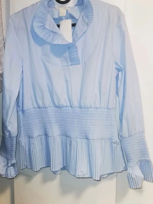 Ljusblå blus med volangkrage - Supersöt ljusblå blus/skjorta med långärmad design och volangdetaljer vid krage, ärmslut och nederkant.  Perfekt för att vara uppklädd. Passar xs