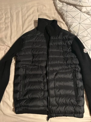 Svart cardigan från Moncler - Säljer en svart pufferjacka från Moncler med quiltad front i dun och stickade ärmar och rygg. Jackan har hög krage, två dragkedjefickor fram och Moncler-logga på vänster ärm. Perfekt för kalla dagar och riktigt snygg till streetwear.