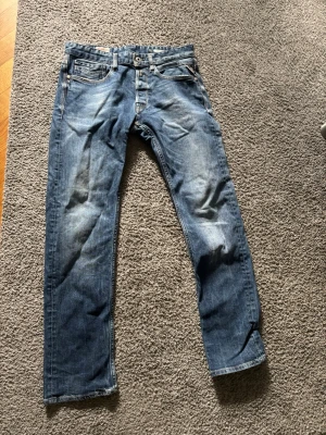 Blå raka jeans från Replay - Säljer ett par blå raka jeans från Replay med snygga slitningar och klassisk femficksmodell. Jeansen har normal midja och detaljerade sömmar. Perfekta för dig som gillar en avslappnad men stilren look.